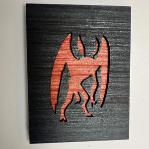 May include: A rectangular black wooden plaque featuring a laser-cut silhouette of a winged creature. The cutout reveals a reddish-brown wood grain texture underneath, creating a high-contrast visual effect against the dark, horizontally grained surface.