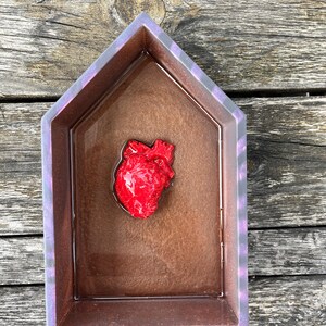 Puede incluir: Una bandeja de resina con forma de casa con un objeto rojo en forma de corazón en su interior. La bandeja tiene un interior marrón y un borde morado y gris. La bandeja está sobre una superficie de madera envejecida.