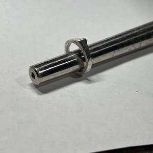 以下が含まれることがあります： 正方形のデザインのシルバーリングが、銀色の棒に取り付けられています。 リングは棒の中央にあり、端には小さな穴があります。 背景は無地の白い表面です。