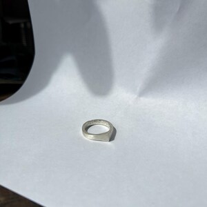 以下が含まれることがあります： 長方形の面を持つシルバーリング。リングは白い背景に置かれています。リングはシンプルでミニマリストなデザインです。光が金属に反射しています。