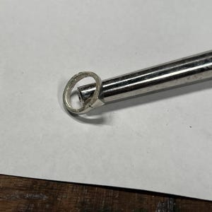 以下が含まれることがあります： 金属工具で加工中の銀の指輪のクローズアップ。 指輪は白い表面にあり、左側にもう一つの指輪が見えます。 ツールは銀色で円筒形です。