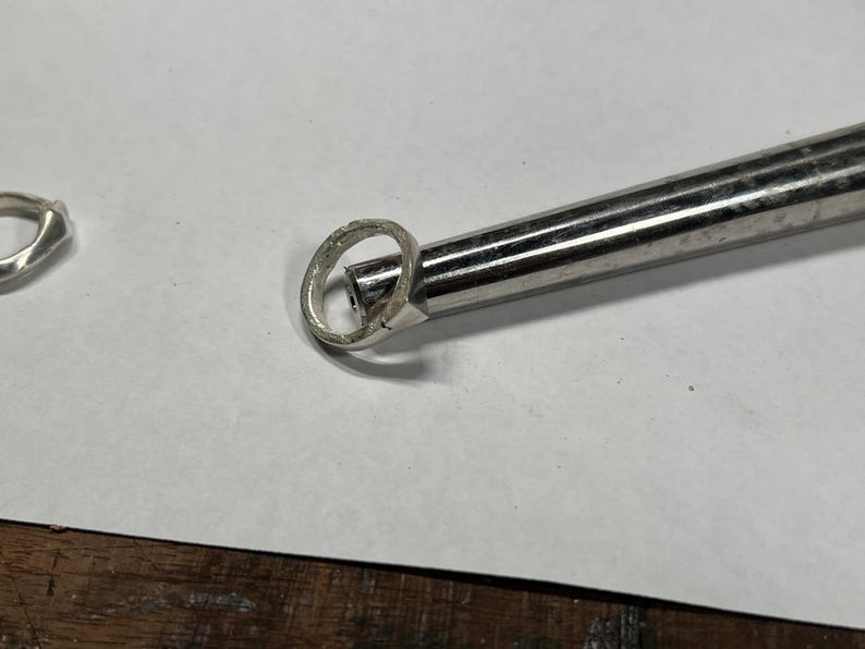 以下が含まれることがあります： 金属工具で加工中の銀の指輪のクローズアップ。 指輪は白い表面にあり、左側にもう一つの指輪が見えます。 ツールは銀色で円筒形です。