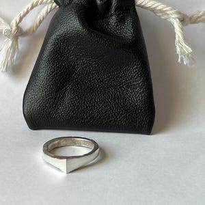 May include: A silver ring with a geometric design, featuring a triangular shape at the front. The ring sits next to a small, black leather drawstring pouch with a white cord closure. The background is a plain, light surface.