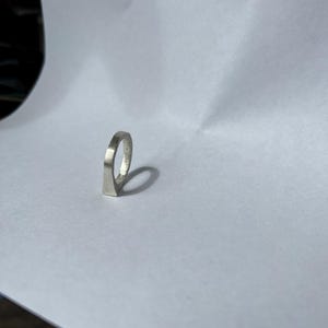以下が含まれることがあります： 幾何学模様のシルバーリング。リングは、ループを形成するために上昇する平らなバンドを持っています。リングは白い背景に配置されています。