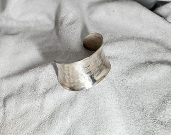 以下が含まれることがあります： 磨き上げられた銀色の金属で作られた幅広のオープンカフブレスレット。ブレスレットの表面には独特のハンマー仕上げの質感が施されており、湾曲した硬いバンド構造全体に小さな反射性のくぼみが連なっています。