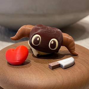 以下が含まれることがあります： 大きな目をした、茶色とタン色の気まぐれな雄羊の置物。赤いハートと小さなナイフが木製の台座に置かれています。台座は、2つの積み重ねられた丸い木片で構成されています。緑の苔が入った大きな灰色のボウルが背景にあります。