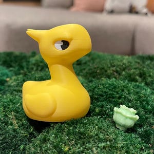 May include: A vibrant yellow duck ornament with a black and white eye, and a small green rose, placed on green moss. The duck has a smooth, rounded design. The background includes a grey sofa and decorative cushions.