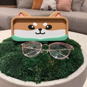 May include: Pink-framed spectacles sit on green moss in a concrete bowl. Above, a brown and white tray with a Shiba Inu dog face design and green trim. The tray has a lid with ears. The moss is in a concrete bowl.