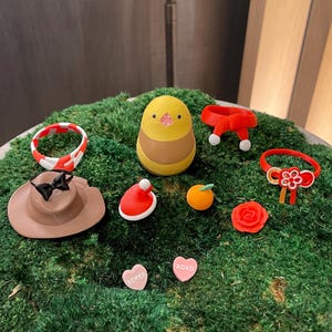 Puede incluir: Una figurita de pájaro amarillo y marrón con una bufanda roja, un gorro de Papá Noel rojo, un sombrero marrón con un lazo negro, una rosa roja, una diadema roja y blanca, un anillo rojo y blanco con un lazo rojo, una naranja pequeña y dos corazones rosas con las palabras "SCREE" y "XOXO" en ellos.