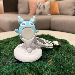 May include: A light blue and white cartoon-style charging stand for a smartwatch. The stand has a round base and a whimsical character design with cloud accents. A white USB cable is connected to the stand.