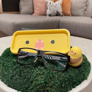 May include: Black rectangular eyeglasses with a yellow case and a small yellow chick-shaped figurine rest on a bed of green moss in a white bowl. The eyeglasses have a black frame and clear lenses.