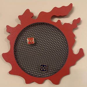 Könnte beinhalten: Eine rote, flammenförmige Wanddekoration mit einem kreisförmigen, grauen Netzmittelpunkt. Zwei kleine, quadratische Anstecknadeln mit farbenfrohen Designs sind am Netz befestigt. Die Anstecknadeln haben rote, goldene und blaue Akzente. Die Dekoration dient wahrscheinlich zum Präsentieren von Anstecknadeln oder kleinen Gegenständen.