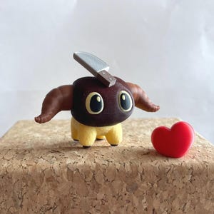 以下が含まれることがあります： 茶色の頭、角、頭にナイフが付いた、気まぐれな3Dプリントの生き物の置物。 生き物には大きくて丸い目と黄色の足があります。 赤いハートが置物の隣にあります。