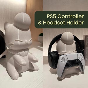 May include: A light grey cat-shaped holder for a PS5 controller and headset. The holder has a round head and a scarf-like detail around the neck. The text "PS5 Controller & Headset Holder" is in a green rectangle.