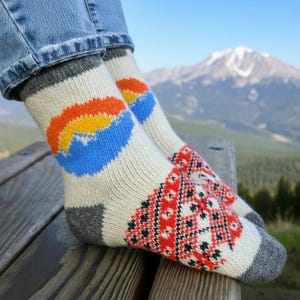 May include: A pair of cream-coloured knitted socks with a mountain design. The socks have a blue, orange, and yellow mountain graphic on the upper part and a red, black, and white geometric pattern on the toes. The cuffs and heels are grey.