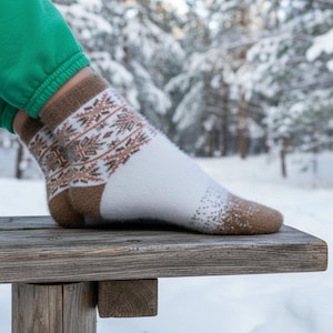 May include: A close-up of a pair of patterned socks. The socks feature a white body, brown heel and toe, and a detailed brown and tan design around the ankle. They are resting on a wooden surface, with a snowy forest visible in the background.