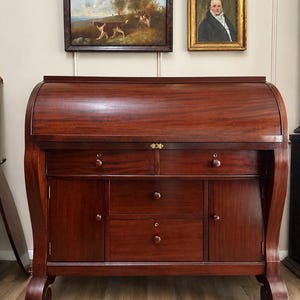 May include: A dark mahogany roll-top desk featuring a curved sliding cover, two upper drawers with round wooden knobs, two side cabinets, and two central drawers. The desk stands on curved legs and includes a small brass keyhole plate.