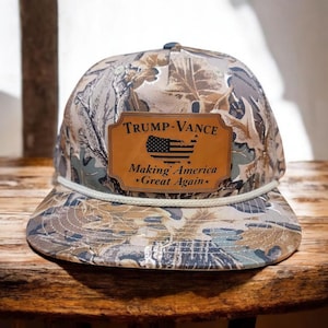 May include: A camouflage baseball cap with a brown leather patch that says "TRUMP-VANCE Making America Great Again" with a black and white American flag inside the patch.