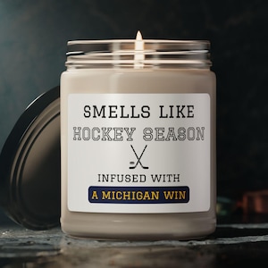 May include: A lit candle in a glass jar with a black lid. The label reads "Smells Like Hockey Season" with crossed hockey sticks and "Infused with a Michigan Win."