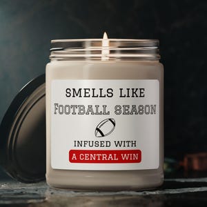 Puede incluir: Una vela encendida en un tarro de cristal con tapa negra. La etiqueta dice "Smells Like Football Season" con un gráfico de fútbol americano y "Infused with A Central Win" en rojo. La vela está sobre una superficie oscura.