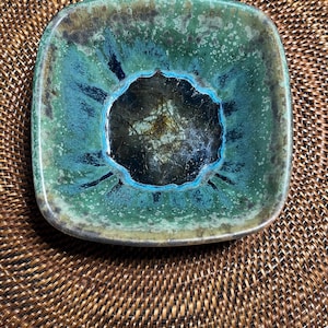 May include: Square ceramic dish with rounded corners. The dish features a teal and green glaze with a dark center. The rim has a darker green color. The dish is sitting on a woven brown mat.