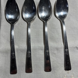 May include: Four silver spoons with oval bowls and long, slender handles. The handles have a decorative design near the base and a rectangular end. The spoons are arranged on a light gray fabric, suggesting a dining or kitchen setting.