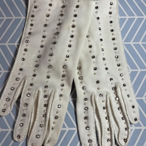May include: A pair of white gloves with rows of silver-toned rhinestones and small white pearls. The gloves are made of a soft, possibly felt-like material. The gloves are laid out on a light blue and white patterned surface.