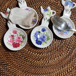 May include: Four white porcelain tea bag holders with floral designs. Each holder has a small teapot-shaped handle. One holder has a tea bag, and another has a silver spoon. The floral patterns include pink, red, and blue roses.