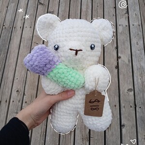 May include: White crocheted teddy bear holding a purple and green crocheted flower. The bear has a brown tag with the text "$18" on it.