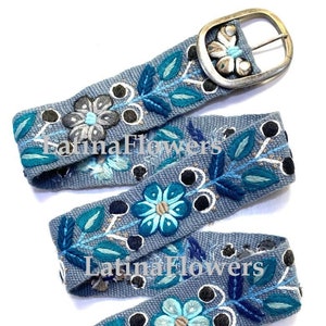 May include: A blue embroidered belt with a silver buckle. The belt is decorated with flowers and leaves in shades of blue, green, and white. The belt is made of a woven fabric and has a slightly textured surface.