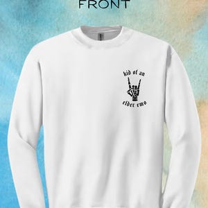 Może przedstawiać: Biała bluza z czarnym nadrukiem graficznym przedstawiającym szkieletową dłoń wykonującą znak rock and roll. Pod ręką nadrukowany jest tekst "kid of an elder emo".