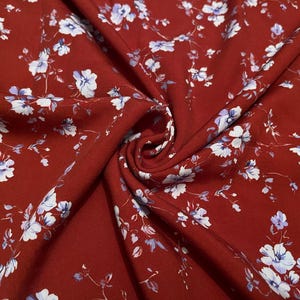 May include: A deep rust-red fabric featuring a repeating pattern of small, delicate white and light blue flowers with thin, dark stems. The material is gathered in the center, creating soft folds and shadows that highlight the floral print.
