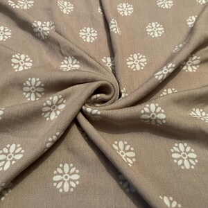 May include: Close-up of a tan fabric with a repeating pattern of white floral designs. The fabric is draped and folded, creating shadows and texture. The floral pattern is evenly spaced across the material.