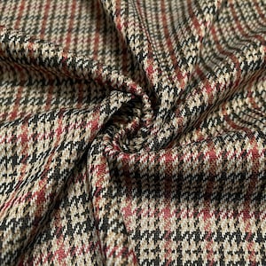 May include: Close-up of a folded fabric with a classic houndstooth pattern. The fabric features a blend of black, beige, and red threads, creating a textured and detailed appearance. The weave is visible, adding depth to the textile.