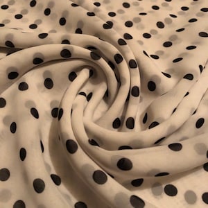 May include: White fabric with a pattern of black polka dots. The fabric is draped and folded, creating soft curves and shadows. The polka dots vary in size and are evenly spaced across the material. The fabric appears to be lightweight and flowing.
