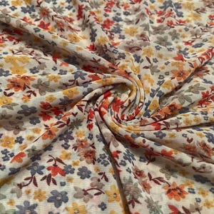 May include: Close-up of a floral fabric with a cream background. The fabric is covered in small flowers in shades of yellow, orange, red, blue, and gray. The fabric is gathered and folded, showing the texture and pattern.