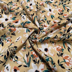 May include: A mustard yellow fabric featuring a repeating floral pattern. The design consists of white, orange, and dark green flowers with dark green leaves and stems scattered across the surface, creating a dense and colorful botanical print.