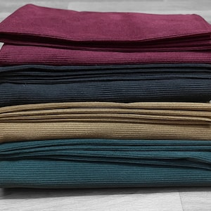 May include: A stack of five fabric swatches in different colors and textures. The swatches are folded and arranged in a pile, with the top swatch being a dark red color. The other swatches are a lighter red, black, brown, and dark green.