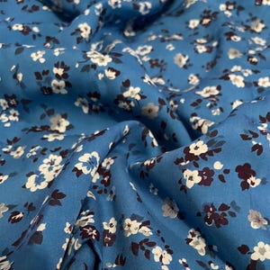 May include: A blue fabric featuring a repeating floral pattern with small white and dark brown blossoms. The material is draped in soft folds, creating a textured surface that highlights the contrast between the blue background and the flowers.