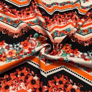 May include: Close-up of a fabric with a vibrant floral and geometric pattern. The design features orange, red, and white flowers set against black and white chevron stripes and orange bands. The fabric is folded, showing its texture.