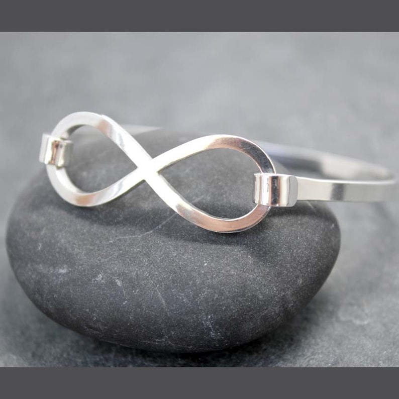 May include: A silver-colored bangle bracelet featuring an infinity symbol design. The bracelet is made of a shiny metal and is displayed on a dark gray stone. The infinity symbol is the focal point, with a sleek, minimalist aesthetic.
