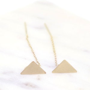 May include: A pair of gold-colored triangle earrings. Each earring features a small, matte gold triangle suspended from a delicate chain. The earrings are displayed on a white surface, creating a minimalist aesthetic.