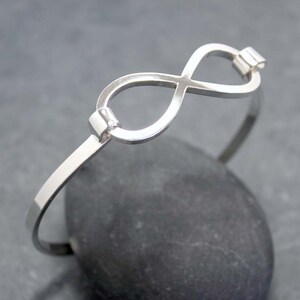 May include: A silver-colored bangle bracelet featuring an infinity symbol at the center. The bracelet has a simple, elegant design with a polished finish. The infinity symbol is connected to the bracelet's band with small, rectangular accents. The bracelet is displayed on a dark gray stone.