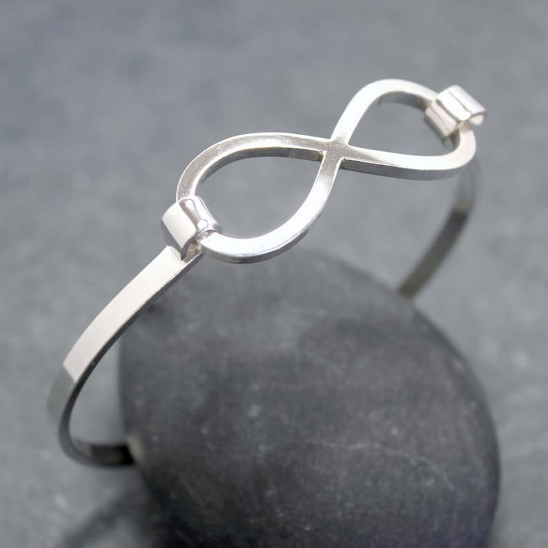 May include: A silver-colored bangle bracelet featuring an infinity symbol at the center. The bracelet has a simple, elegant design with a polished finish. The infinity symbol is connected to the bracelet's band with small, rectangular accents. The bracelet is displayed on a dark gray stone.