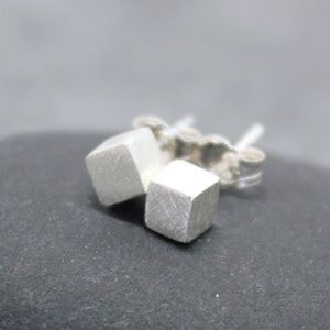 May include: A pair of silver cube stud earrings. The earrings are square-shaped with a brushed metal finish. The earring posts and backs are also visible. The earrings are displayed on a dark gray surface.