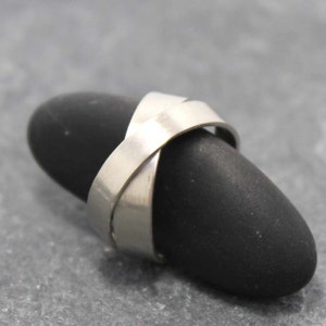 Peut inclure: Une bague argentée présentant une finition en métal brossé lisse avec un design croisé où deux bandes s'entrecroisent au centre. La bague possède une forme fluide et continue avec un aspect mat, offrant une esthétique simple et épurée.