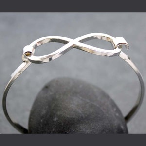 May include: A silver-colored bangle bracelet featuring an infinity symbol design. The bracelet is made of a thin, polished metal and has a simple, elegant style. The infinity symbol is the focal point, adding a touch of symbolism to the jewelry.