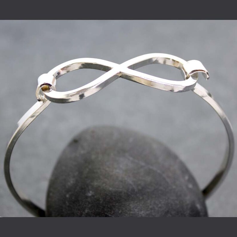 May include: A silver-colored bangle bracelet featuring an infinity symbol design. The bracelet is made of a thin, polished metal and has a simple, elegant style. The infinity symbol is the focal point, adding a touch of symbolism to the jewelry.