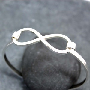 May include: A silver-colored bangle bracelet featuring an infinity symbol design. The bracelet is made of a thin, polished metal and has a simple, elegant style. The infinity symbol is the focal point, adding a touch of symbolism to the jewelry.