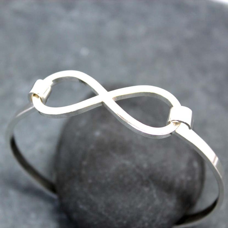 May include: A silver-colored bangle bracelet featuring an infinity symbol design. The bracelet is made of a thin, polished metal and has a simple, elegant style. The infinity symbol is the focal point, adding a touch of symbolism to the jewelry.